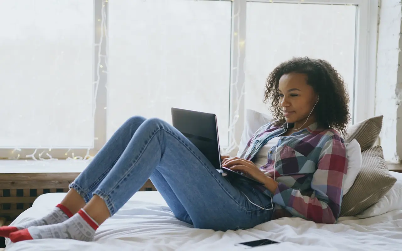 A student studying online from bed, laptop and earbuds, string lights behind her